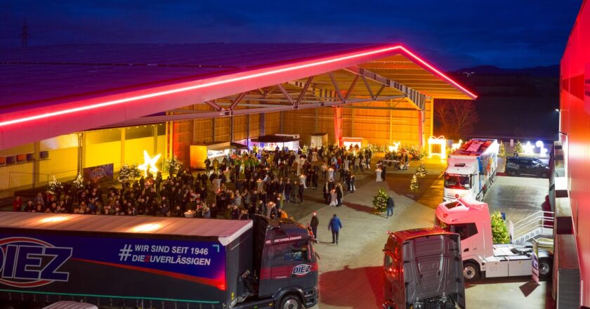 A large open-air transport depot hosting a night-time launch event, with trucks parked in front of a crowd beneath a roof lit with red LED lighting.